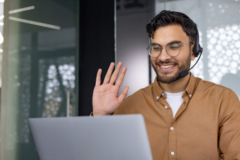 Smiling man with glasses and headset waving during a video call on his laptop, in a modern office setting with glass walls.