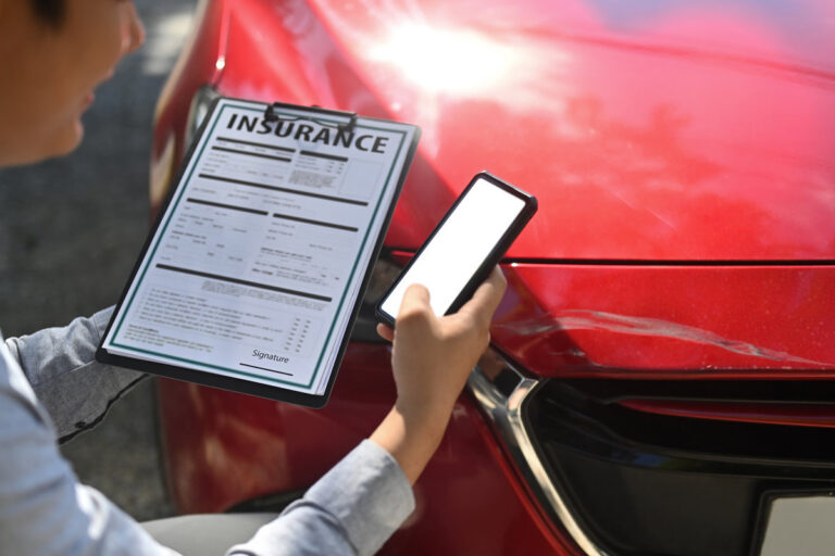 Insurance agent holding insurance claim form and using smart phone during inspecting damaged car.